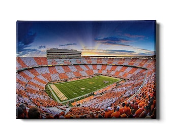 May include: A panoramic view of a packed football stadium with orange and white seats. The field is green with white yard lines and a brown checkerboard pattern in the end zone. The sky is blue with a few clouds and a sunset in the distance.