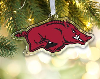 Op de afbeelding: Een doorzichtig acryl kerstornament in de vorm van een aanvallend wild zwijn. Het zwijn is dieprood gekleurd met zwarte omlijningen en witte slagtanden. Een wit lint is aan de bovenkant bevestigd en een klein geregistreerd handelsmerksymbool is zichtbaar.