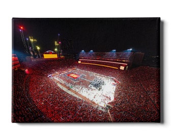 May include: A night time aerial view of a packed football stadium with a lit up field and a large crowd of people wearing red. The stadium is lit up with lights and there are fireworks in the sky.