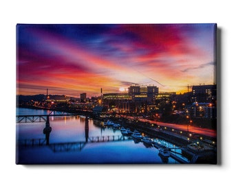 May include: A panoramic view of a city skyline at dusk with a bridge over a river. The sky is a vibrant mix of pink, purple, and orange with clouds. The city lights are reflected in the water.