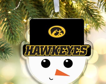 May include: A clear, snowman-shaped ornament with a black hat featuring the Iowa Hawkeyes logo and the word "HAWKEYES" in gold lettering. The snowman has black eyes, an orange carrot nose, and a curved mouth. A white ribbon is attached for hanging.