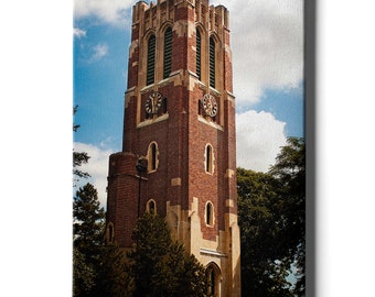 May include: A tall, brick tower with two clock faces and arched windows. The tower is surrounded by green trees and a blue sky with white clouds.