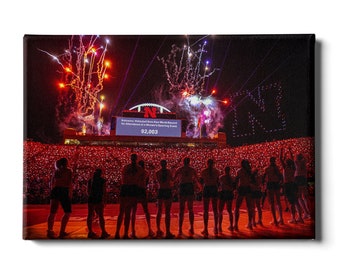 May include: A large crowd of people in a stadium with fireworks exploding in the night sky. The crowd is holding up red glow sticks, and a large scoreboard displays the text "Nebraska Volleyball Sets New World Record for Attendance at a Men's Sporting Event 92,003".