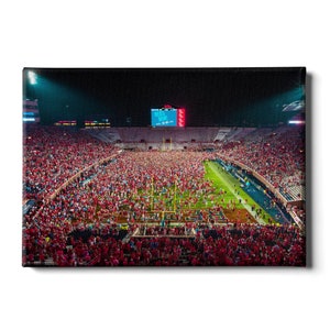 May include: A night time view of a packed football stadium with a large scoreboard displaying "REBS WIN" in blue text. The stadium is lit by bright lights and the crowd is wearing red.