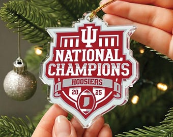 May include: A clear, shield-shaped Christmas ornament with a red and white design. The ornament features the text "NATIONAL CHAMPIONS HOOSIERS 2025" and the Indiana state outline. It is suspended by a gold string.