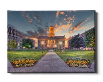 May include: A large, beige building with a dome and columns, illuminated by warm lights, stands on a grassy lawn with a paved walkway leading to the front entrance. The sky is a vibrant blue with pink and orange clouds.