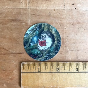 Op de afbeelding: Een ronde sticker met een cartoon-egel die een bril draagt en een boek leest bij kaarslicht in een boomhol. De egel wordt omringd door paddenstoelen en andere boswezens.