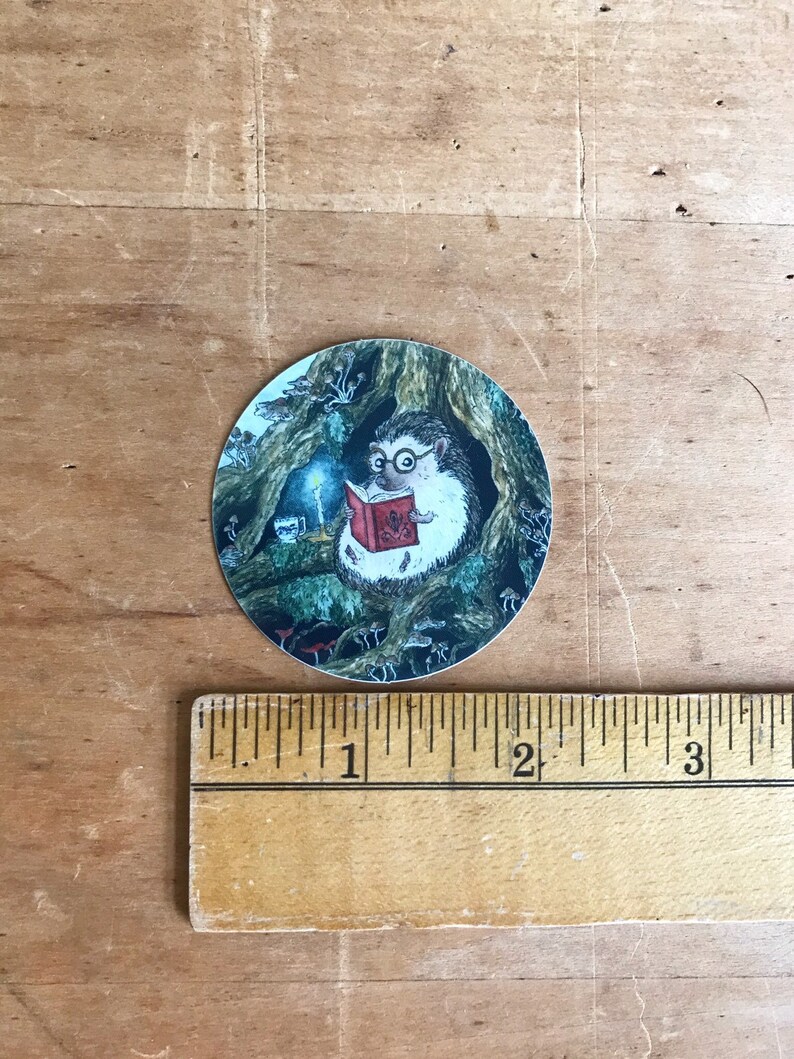 Op de afbeelding: Een ronde sticker met een cartoon-egel die een bril draagt en een boek leest bij kaarslicht in een boomhol. De egel wordt omringd door paddenstoelen en andere boswezens.