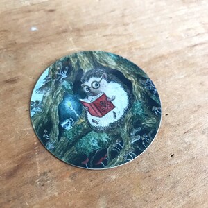 Op de afbeelding: Een ronde sticker met een grillige illustratie van een egel die een bril draagt en een boek leest bij kaarslicht in een holle boom. De sc&egrave;ne is omringd door paddenstoelen.