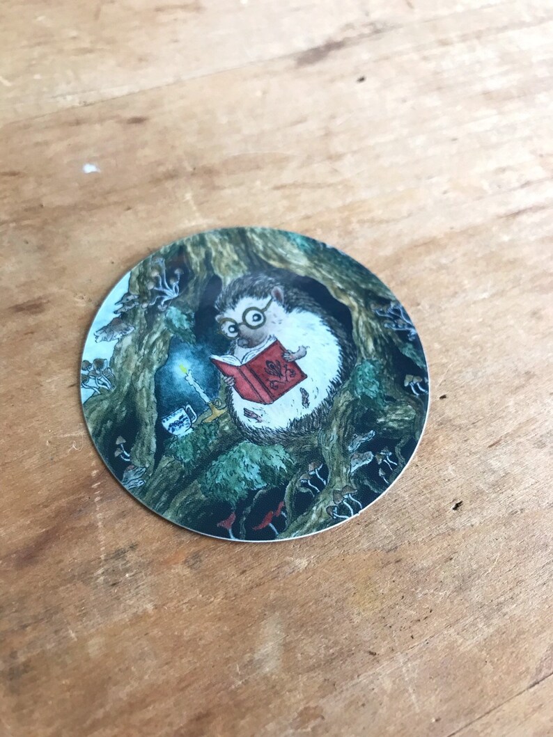 Op de afbeelding: Een ronde sticker met een grillige illustratie van een egel die een bril draagt en een boek leest bij kaarslicht in een holle boom. De sc&egrave;ne is omringd door paddenstoelen.