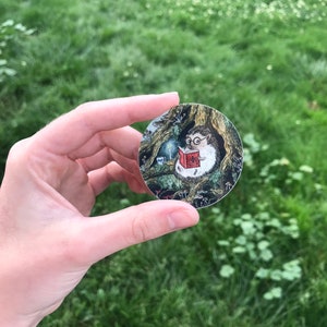 Op de afbeelding: Een ronde sticker met een cartoon-egel die een bril draagt en een boek leest in een bossetting. De egel zit op een boomstronk met paddenstoelen en andere bosselementen eromheen.