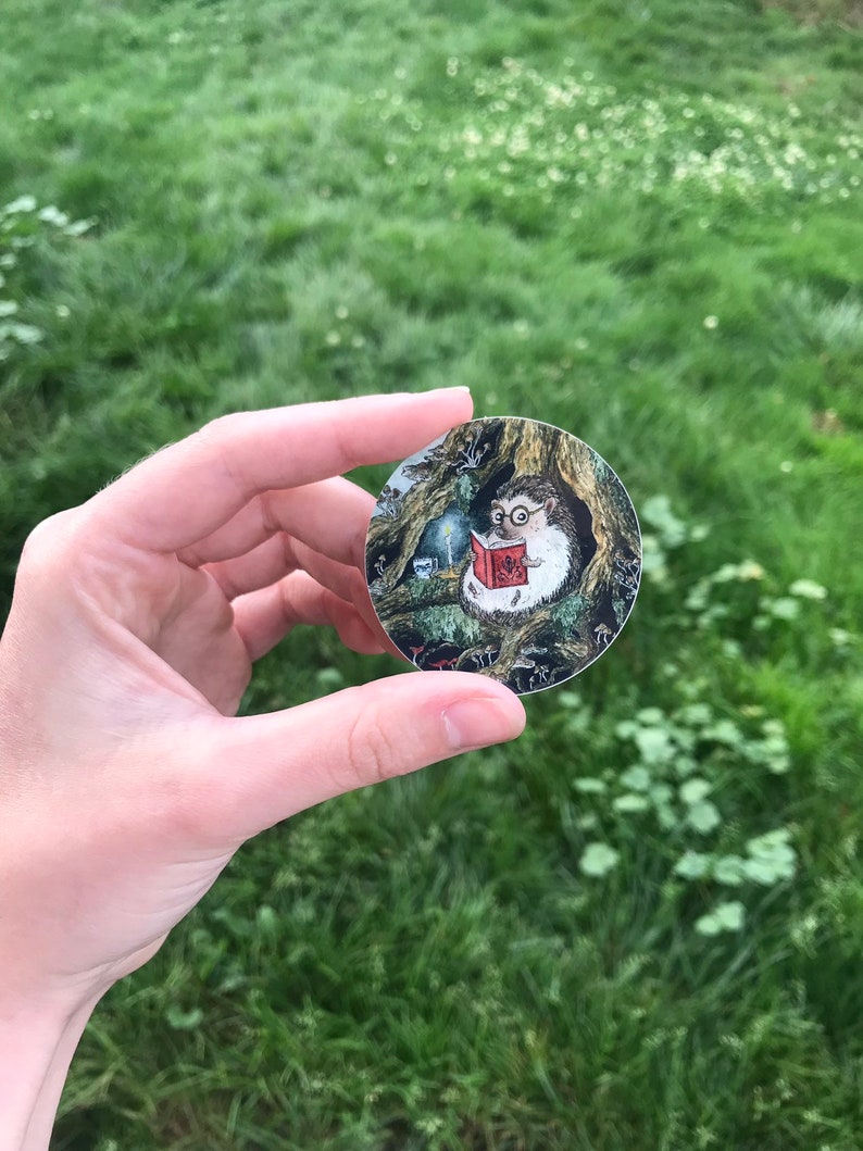 Op de afbeelding: Een ronde sticker met een cartoon-egel die een bril draagt en een boek leest in een bossetting. De egel zit op een boomstronk met paddenstoelen en andere bosselementen eromheen.