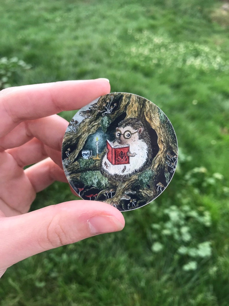 Op de afbeelding: Een ronde sticker met een egel die een bril draagt en een boek leest in een bossetting. De egel zit op een tak met paddenstoelen en andere bosselementen eromheen.