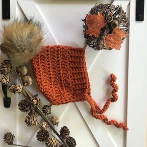 May include: A crocheted baby bonnet in a burnt orange colour with a fluffy pom pom on top. The bonnet has a long tie that is also crocheted.