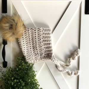 May include: A beige crocheted baby bonnet with a brown faux fur pom pom and a long tie. The bonnet is on a white background.