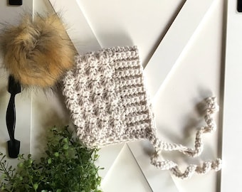 Peut inclure: Un bonnet de bébé en crochet avec un motif texturé et un pompon moelleux sur le dessus. Le bonnet est attaché avec une longue ficelle en crochet.