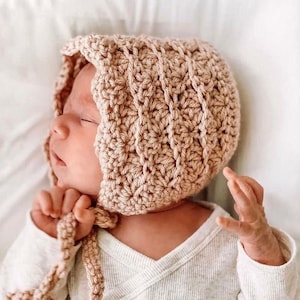 May include: A soft, light brown crocheted baby bonnet with a tie. The bonnet has a textured stitch pattern and is tied under the chin.