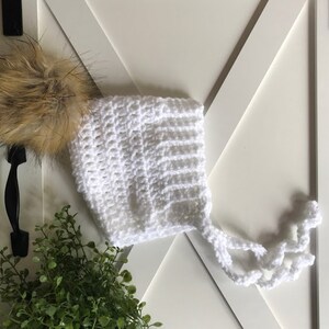 May include: White crocheted baby bonnet with a fluffy brown pom pom. The bonnet has a long tie that can be tied under the chin.
