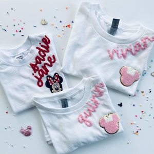 Pode incluir: Três camisetas brancas com nomes personalizados bordados em fio rosa e vermelho. Uma camisa apresenta um patch da Minnie Mouse. As camisas são decoradas com confetes coloridos e enfeites em forma de Mickey Mouse.