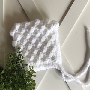 May include: White crocheted baby bonnet with a textured bobble stitch pattern and two long ties.