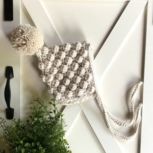 May include: A cream-colored crocheted baby hat with a white pom-pom on top. The hat has a tie at the bottom.