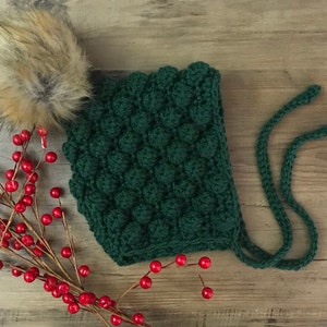 May include: A green crocheted baby bonnet with a fluffy brown pom pom. The bonnet has a tie at the back.