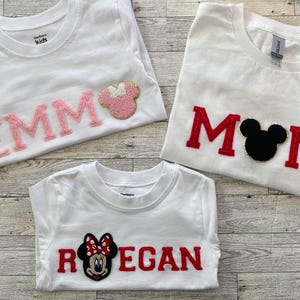 May include: Three white t-shirts with different designs. One shirt has the name "EMM" in pink letters with a pink mouse-shaped design. Another says "MOM" in red letters with a black mouse head. The last shirt says "REGAN" with a Minnie Mouse design.