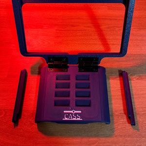 以下が含まれることがあります： 透明なプラスチックフレームが付いた黒いプラスチック製のゲームカートリッジホルダー。ホルダーにはゲームカートリッジ用のスロットが8つあり、前面に「CASS」という文字が印刷されています。