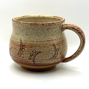Peut inclure: Une tasse en céramique avec un corps arrondi et une anse incurvée. L'extérieur présente un émail crème et beige moucheté avec une série de motifs sculptés en forme de feuilles entourant la base. Le bord est lisse et la base est en argile brune.
