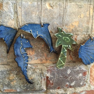 May include: Five ceramic ornaments hanging from twine. The ornaments are shaped like a seahorse, a dolphin, a bird, a palm tree, and a seashell. The ornaments are painted in shades of blue and green.