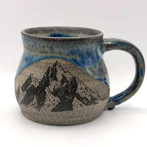 May include: A ceramic mug with a speckled grey base and a mountain range illustration in black ink. The upper portion of the mug features a glossy blue glaze that drips down towards the textured, earthy grey body. It has a curved handle.
