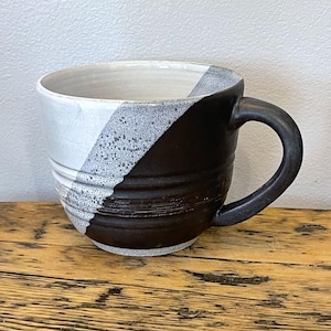 Può includere: Una tazza in ceramica fatta a mano con un design unico. La tazza presenta un motivo a blocchi di colore diagonali, con bianco da un lato, una sezione grigia macchiettata e marrone scuro dall'altro. La tazza ha una forma arrotondata e un manico robusto.