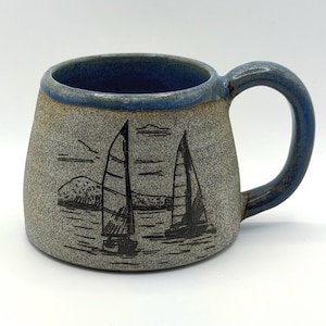 May include: A ceramic mug with a speckled grey body and a blue rim and handle. The exterior features a black line art illustration of two sailboats on the water with a mountain in the background. The mug has a wide base and a curved handle.