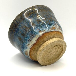 Peut inclure: Une tasse en céramique avec une base beige mouchetée et un émail réactif passant d'une bande ambrée chaude à un motif de gouttes bleu profond et blanc. Le fond de la tasse présente une surface en argile texturée non émaillée avec une marque.