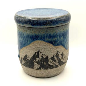 Peut inclure: Un pot en céramique cylindrique avec un couvercle assorti, présentant une base gris clair mouchetée et une glaçure bleu foncé coulant du bord supérieur. L'extérieur affiche une illustration à l'encre noire d'une chaîne de montagnes.