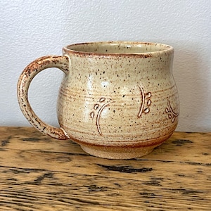 Könnte beinhalten: Eine handgefertigte Keramiktasse mit einer gesprenkelten beigefarbenen Glasur und einem braunen Henkel. Die Tasse hat ein dekoratives Blattdesign und eine breite, abgerundete Form. Die Tasse steht auf einer Holzoberfläche.