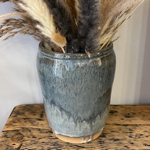 Puede incluir: Un jarrón de cerámica esmaltada azul y gris con boca ancha, que contiene hierbas ornamentales secas. El jarrón tiene una superficie texturizada y una base marrón claro. El jarrón está sobre una mesa de madera rústica.