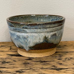 May include: A handcrafted ceramic bowl with a blue and brown glaze. The bowl is round with a wide opening and rests on a small, round, unglazed base. The interior and exterior of the bowl have a textured appearance, suggesting a rustic aesthetic.