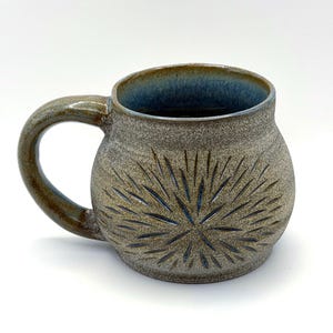 May include: A round ceramic mug with a speckled grey and brown exterior featuring a carved starburst pattern. The interior of the mug is glazed in a deep blue, and the handle is curved and finished in a matching speckled brown glaze.
