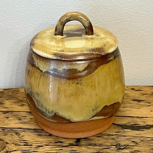 Könnte beinhalten: Ein handgefertigtes Keramikgefäß mit Deckel. Das Gefäß hat eine runde Form mit einem Griff oben. Die Farben sind erdig, mit Brauntönen, Beige und einem Hauch von Gelb. Das Gefäß steht auf einer Holzoberfläche.