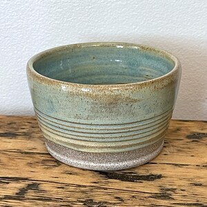 Può includere: Una ciotola in ceramica fatta a mano con interno azzurro chiaro ed esterno strutturato. La ciotola presenta linee orizzontali e una base macchiettata marrone chiaro. Il bordo è leggermente irregolare, aggiungendo al suo fascino artigianale unico.