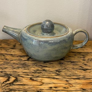 May include: A ceramic teapot with a blue and green glaze. The teapot has a rounded body, a curved spout, and a handle. The lid is topped with a small, round knob. The teapot is sitting on a wooden surface.
