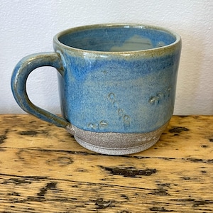 Puede incluir: Una taza de cerámica hecha a mano con un esmalte azul y una base marrón. La taza tiene un asa curva y una superficie texturizada. La taza está sobre una superficie de madera.