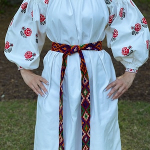 May include: White linen dress with embroidered red roses on the sleeves and a colorful woven belt.