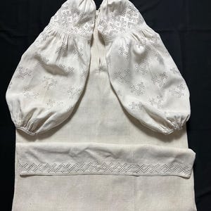 May include: A cream-colored, long-sleeved linen shirt with intricate embroidered details. The sleeves are voluminous, gathered at the cuffs, and feature openwork patterns. The shirt's neckline is simple, and the hemline also has an embroidered border.