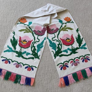 May include: A cream-colored runner featuring a cross-stitch floral pattern in shades of pink, red, orange, and green. The design includes large stylized flowers and smaller blossoms, finished with a multi-colored fringe in blue, pink, and green.