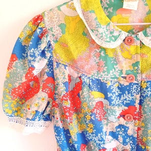 Op de afbeelding: Een blouse met korte mouwen en een levendig bloemenpatroon in blauw, rood, geel en groen. Het kledingstuk heeft een Peter Pan-kraag afgezet met witte kant, pofmouwen met kanten boorden en een sluiting aan de voorzijde met roze knopen.