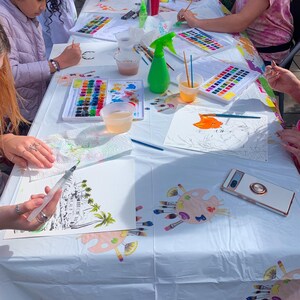 May include: A white table covered in a white tablecloth with paint palettes, paintbrushes, water cups, and art supplies.  Several children are painting on paper at the table.