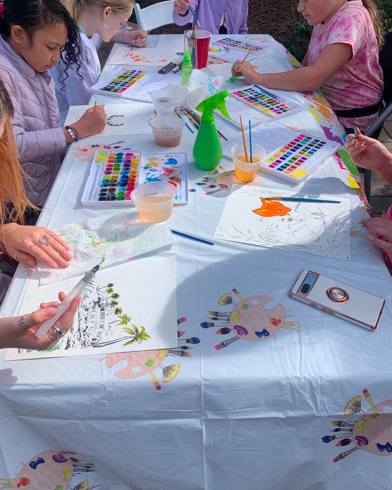 May include: A white table covered in a white tablecloth with paint palettes, paintbrushes, water cups, and art supplies.  Several children are painting on paper at the table.