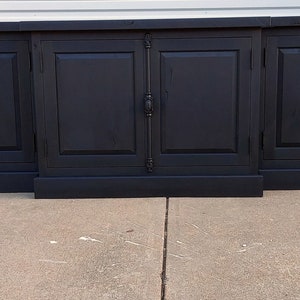 May include: A black wooden cabinet with six doors and black metal hardware. The cabinet has a base and sits on a concrete floor.
